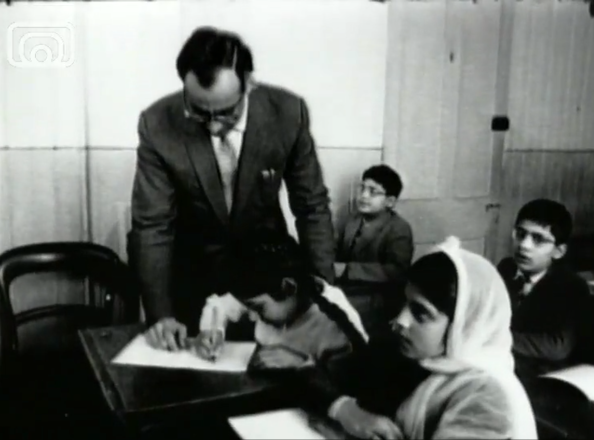 Black and white image, classroom with teacher standing beside pupils